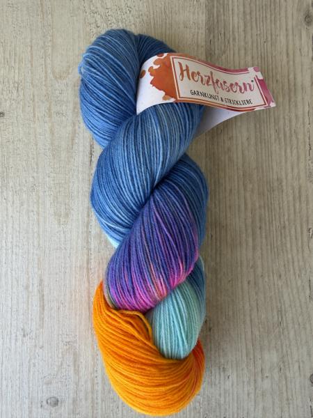 Ein handgefärbter Garnstrang in kräftigem Blau mit Farbverläufen in Orange, Türkis und Pink liegt auf einem hellen Holzuntergrund. Das Garn ist mit einer Herzfasern-Banderole versehen.